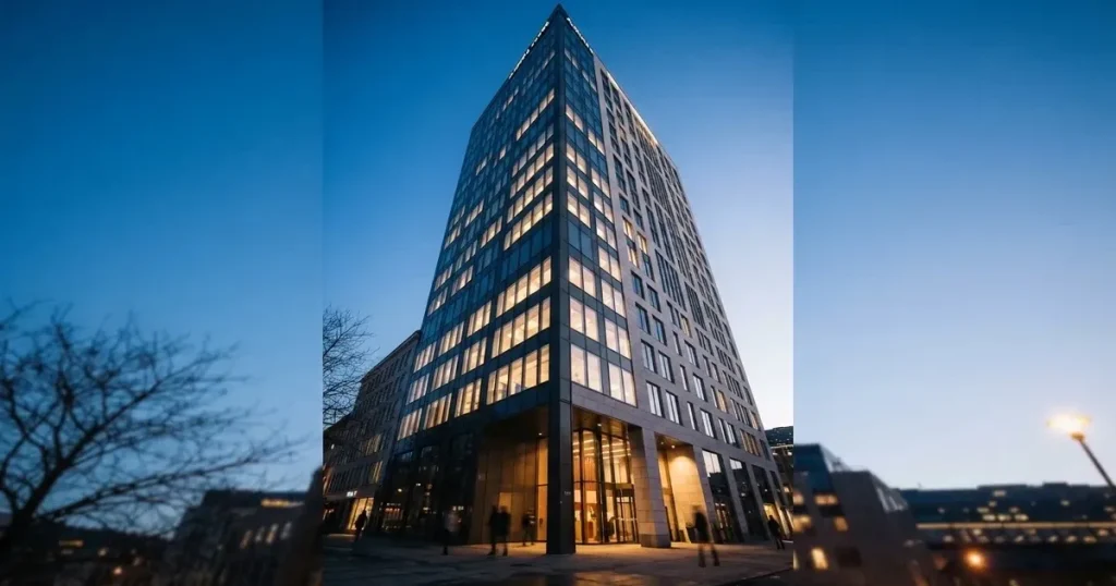 En modern kontorsbyggnad fotograferad underifrån i skymning med tända fönster mot en klarblå himmel.